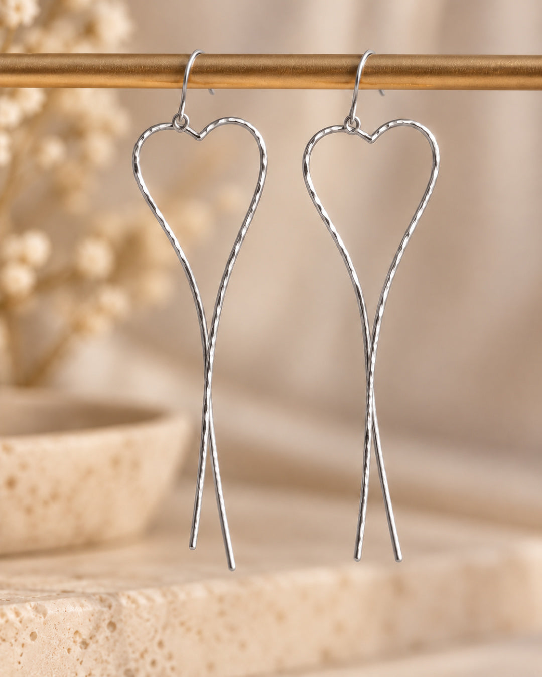 Pair of Allure Heart Earrings in hammered Argentium silver suspended against a warm neutral background, highlighting their open-heart design and long, flowing crossed ends.