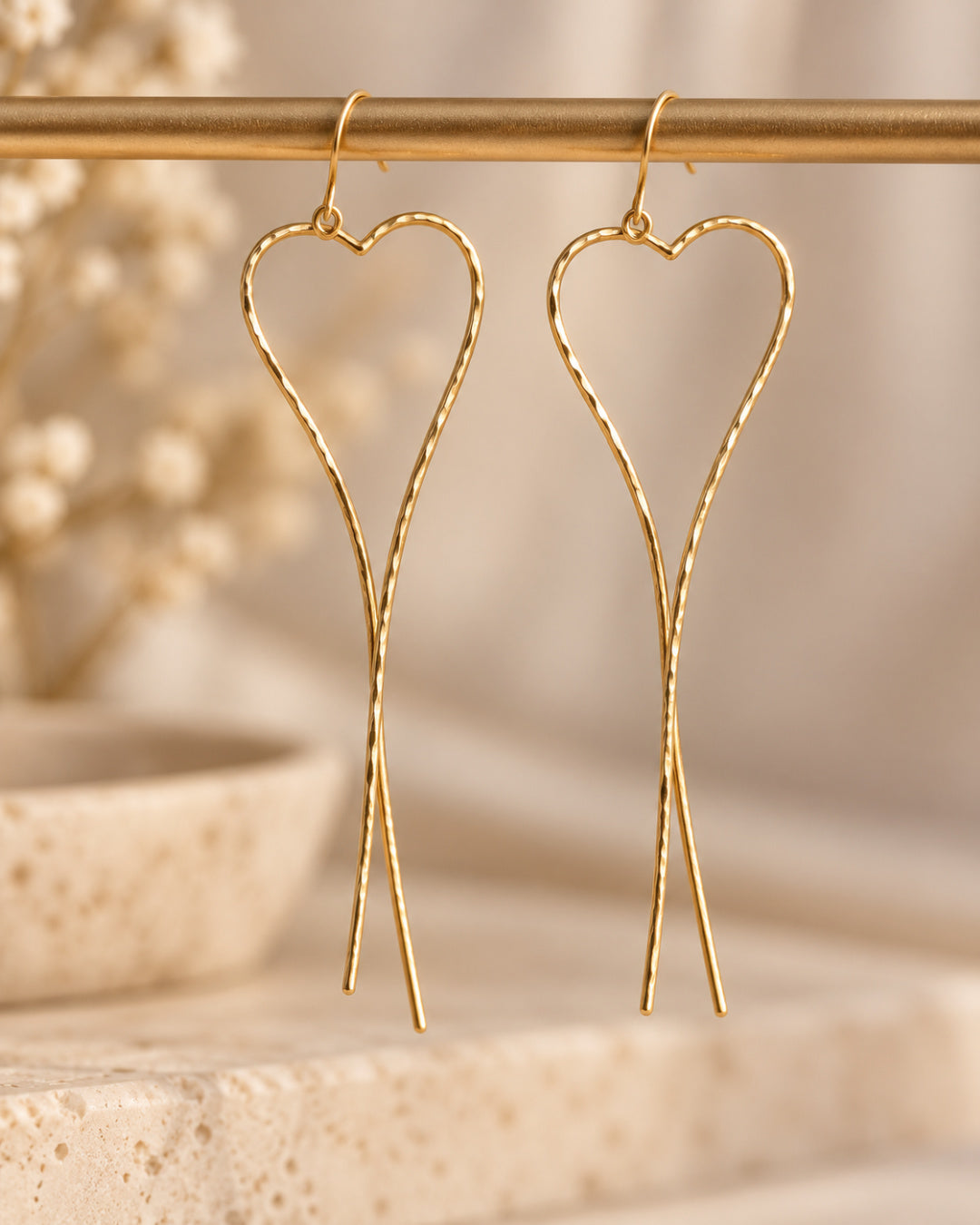 Pair of gold-filled Allure Heart Earrings suspended from a brass rod, showcasing sculptural heart outlines, delicate hammered texture, and elongated crossed wire ends.