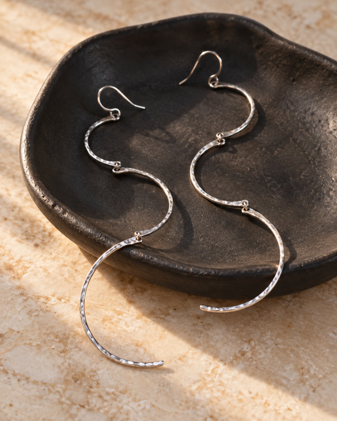 Long hammered silver Cadence Earrings resting across a matte-black ceramic jewelry dish on warm stone.