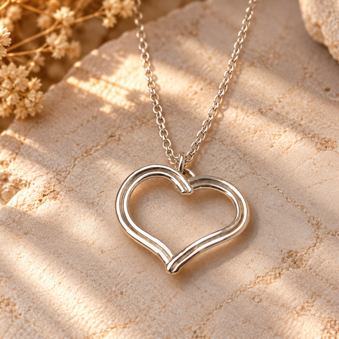 Devotion Heart Necklace in polished Argentium silver displayed on warm cream stone, highlighting its open-heart silhouette, flowing double-line design, and silver rolo chain.