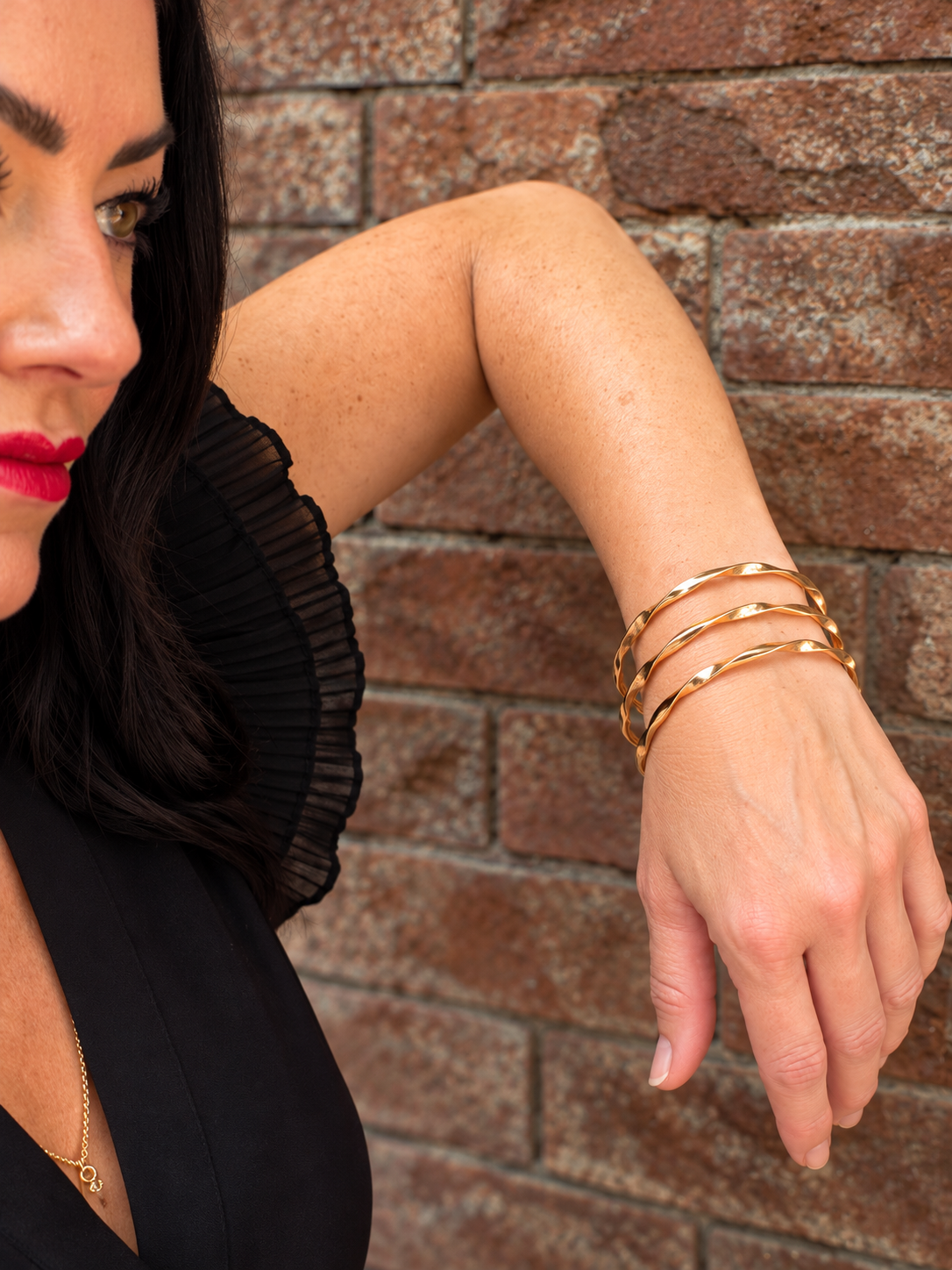 A close-up lifestyle shot of a woman wearing three stacked, polished gold bangle bracelets on her wrist, posed against a rustic brown brick wall.