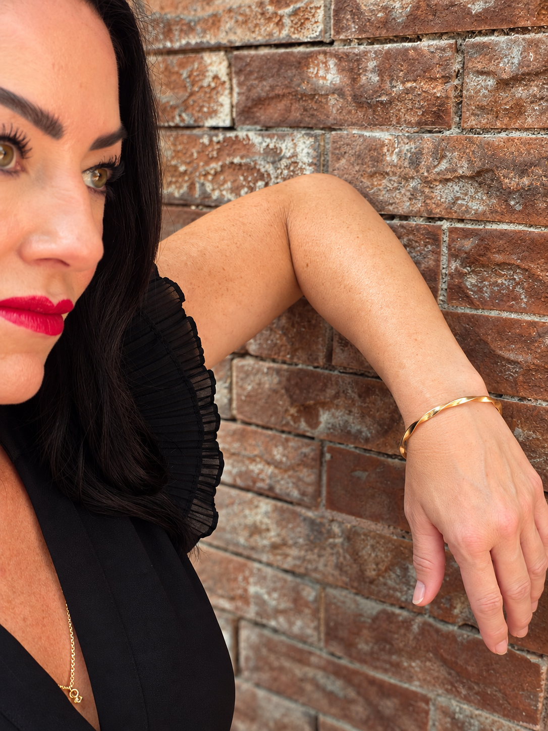 A woman with dark hair and red lipstick leans against a rustic brick wall, wearing a single gold bangle bracelet on her wrist.