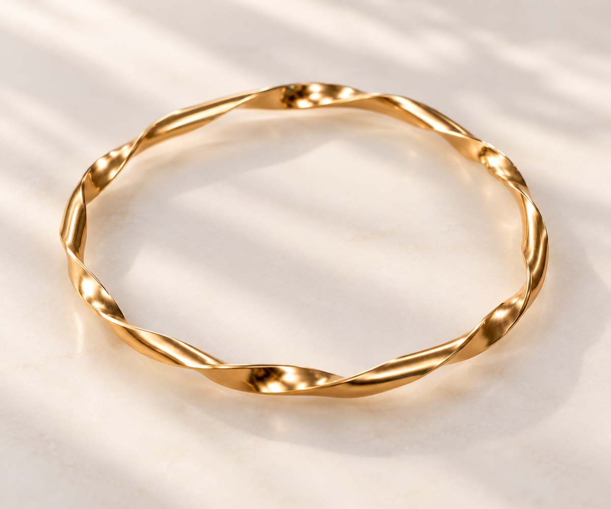 A twisted gold bangle bracelet with a polished finish, displayed on a light beige marble surface with soft leaf shadows.