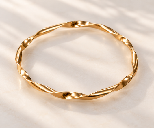 A twisted gold bangle bracelet with a polished finish, displayed on a light beige marble surface with soft leaf shadows.