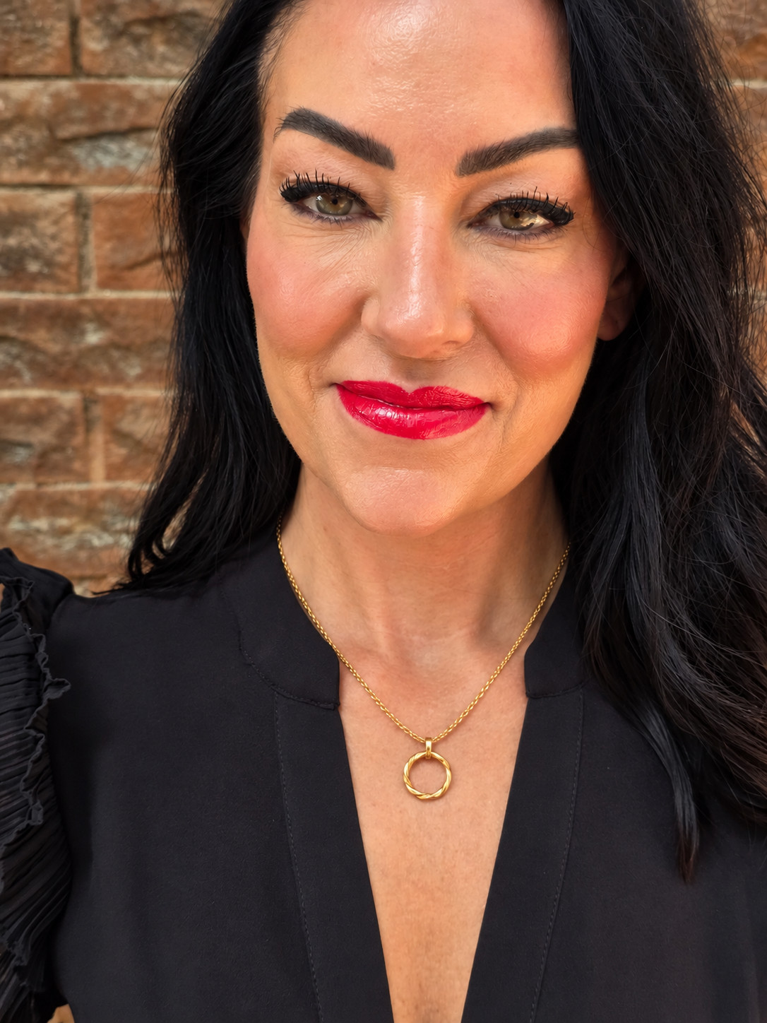 A close-up portrait of a woman with dark hair wearing a delicate gold chain necklace with a simple circular pendant, paired with a black top, set against a rustic brick wall background.