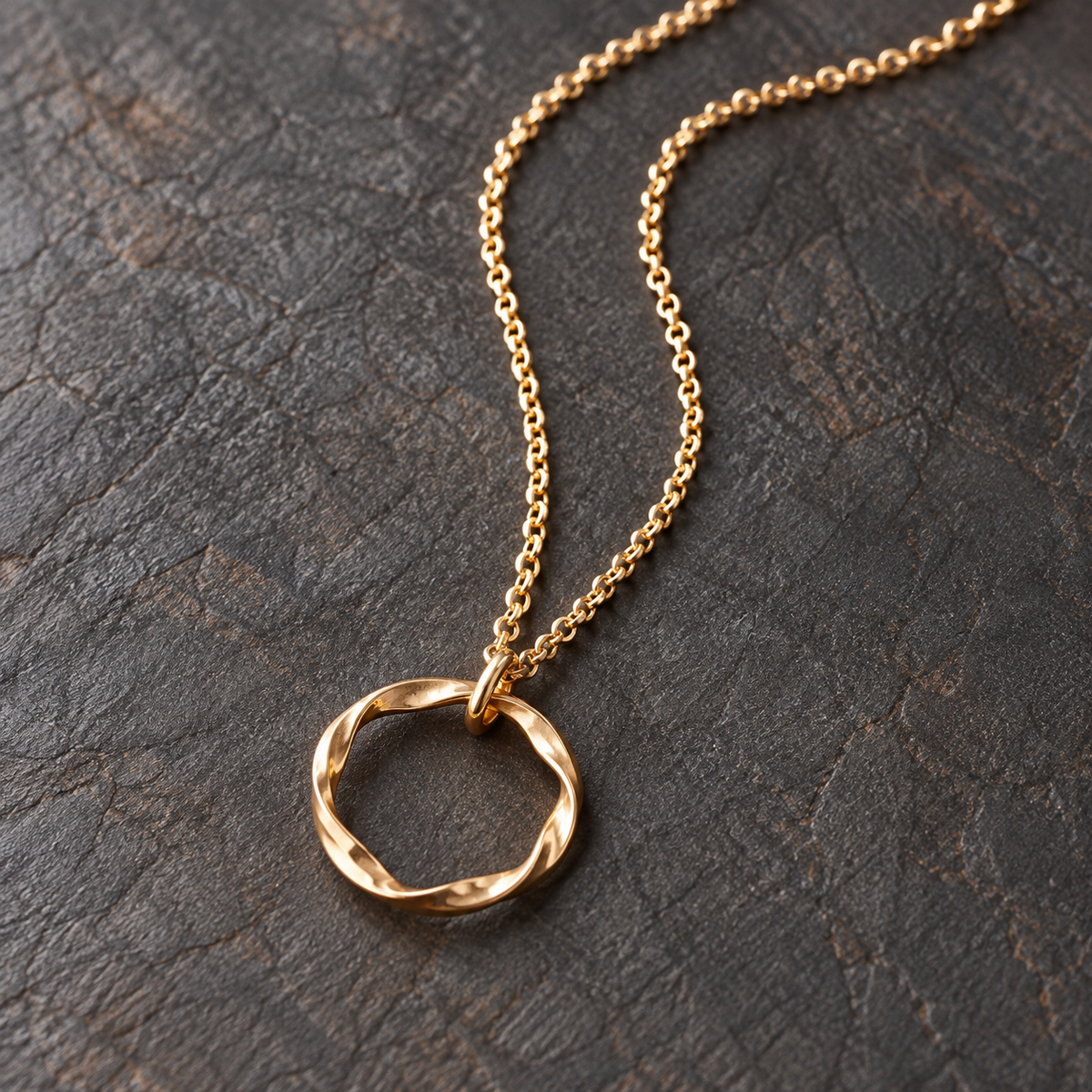 A gold necklace with a twisted circle pendant, displayed on a dark textured leather surface.