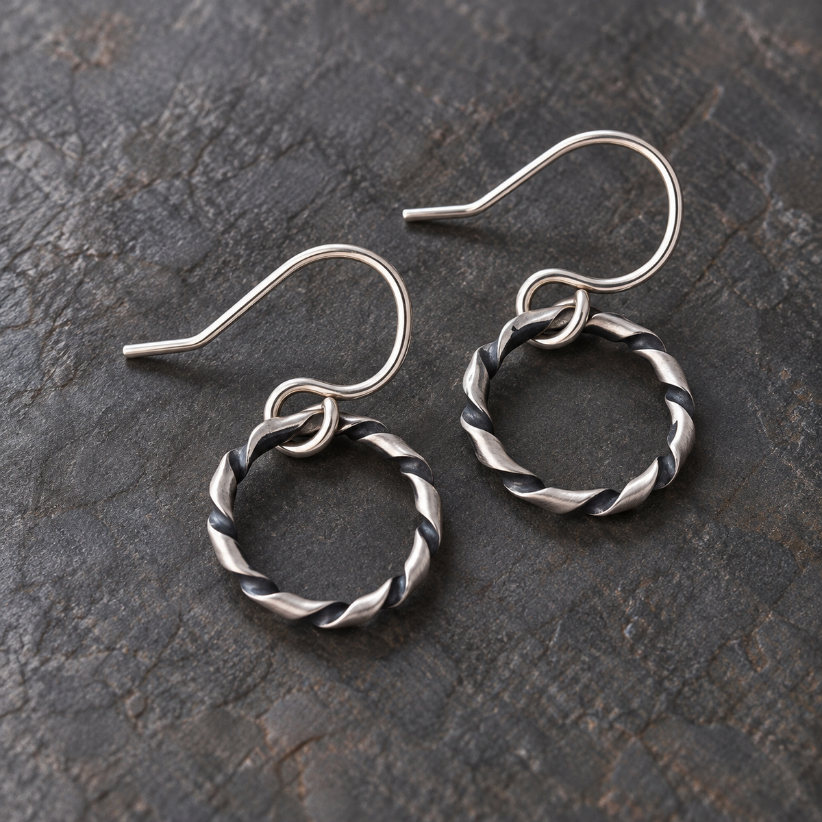 A pair of structural, oxidized argentium silver twisted hoop dangle earrings with fishhook backs, displayed on a dark textured slate surface.