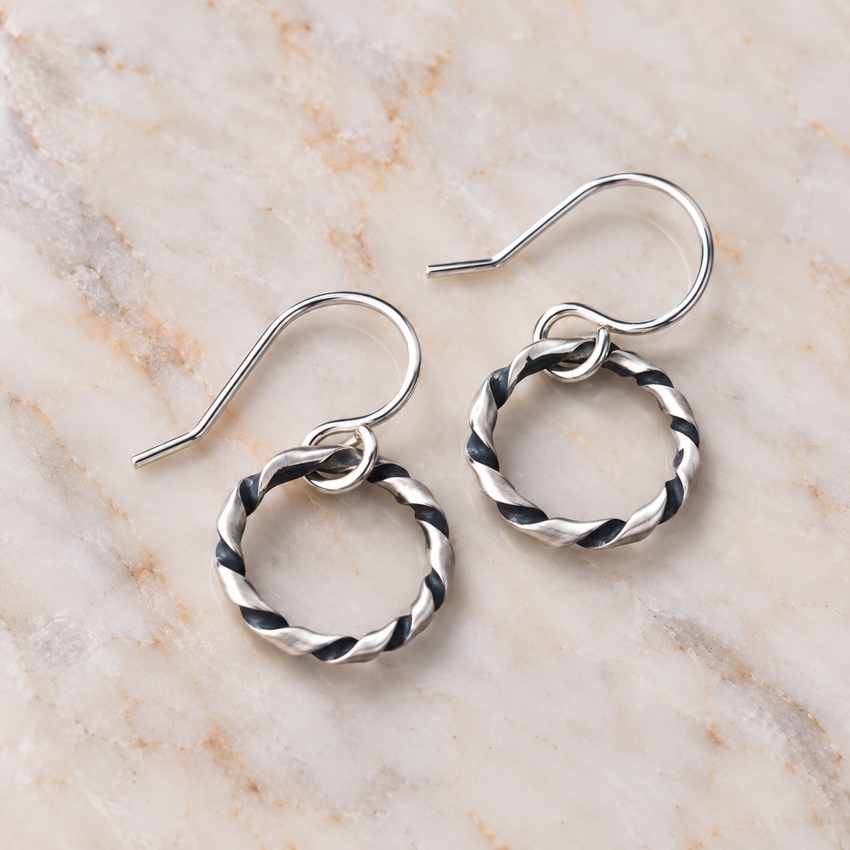 A pair of oxidized argentium silver earrings with twisted circular hoops, displayed on a light beige marble surface.