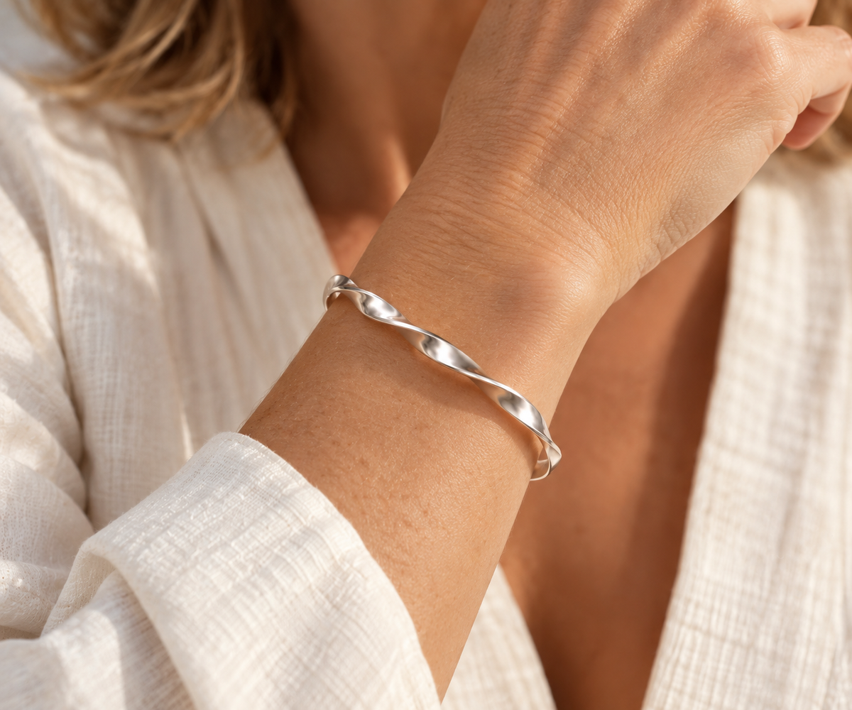 A close-up of a woman's wrist wearing a polished silver twist bangle bracelet, dressed in a textured white linen top.