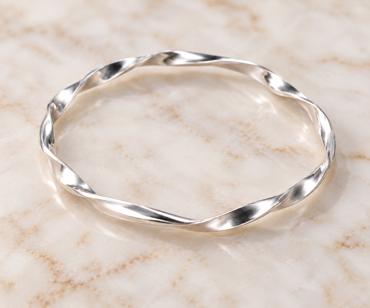 A polished silver bangle bracelet with a twisted, fluid wave design, resting on a light beige marble surface.
