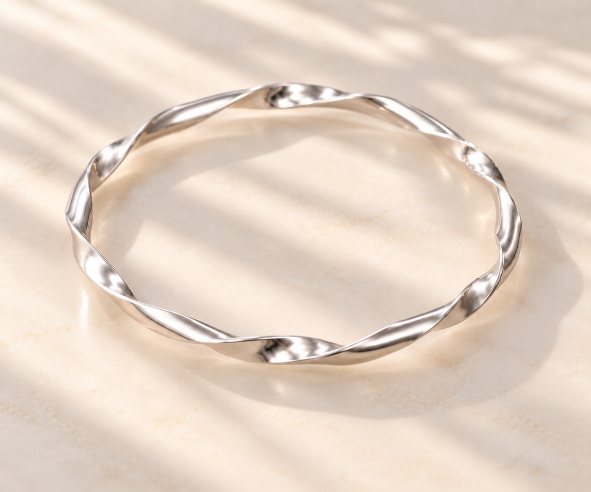 A polished silver twisted bangle bracelet resting on a light beige marble surface with soft, dappled shadows.