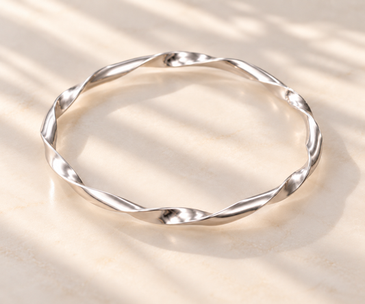 A polished silver twisted bangle bracelet resting on a light beige marble surface with soft, dappled shadows.