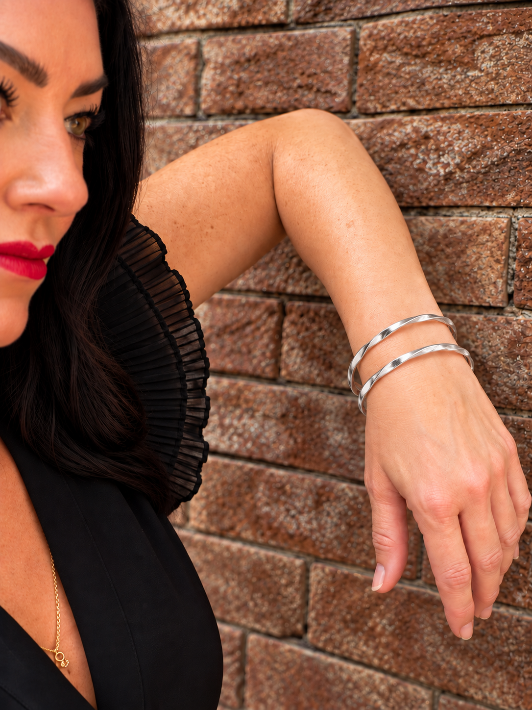 A close-up lifestyle shot of a woman wearing a stack of two polished silver bangle bracelets, leaning against a rustic brown brick wall.