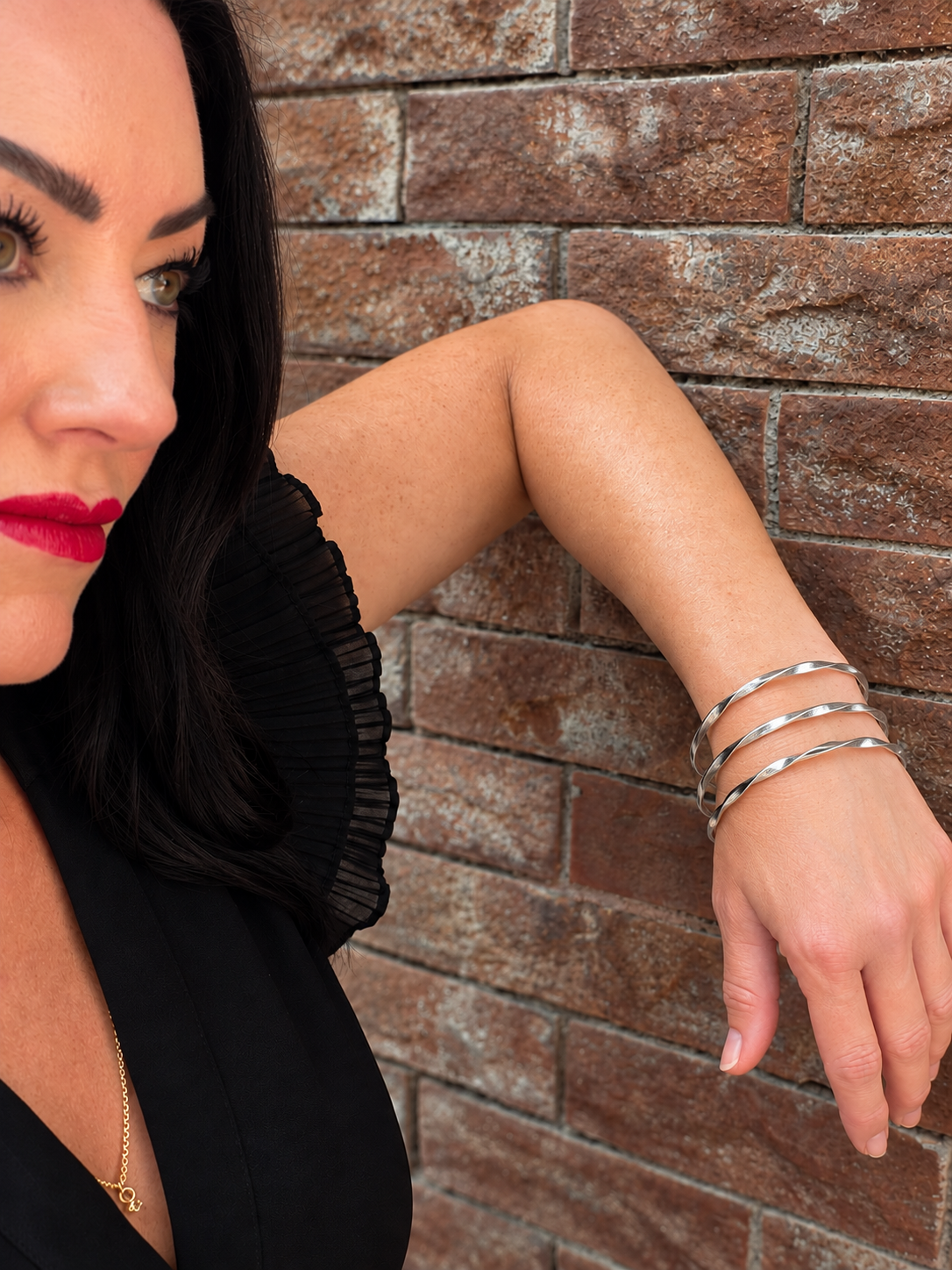A close-up lifestyle shot of a woman with dark hair and red lipstick, wearing a stack of three sculptural silver bangle bracelets on her wrist, leaning against a rustic brown brick wall.