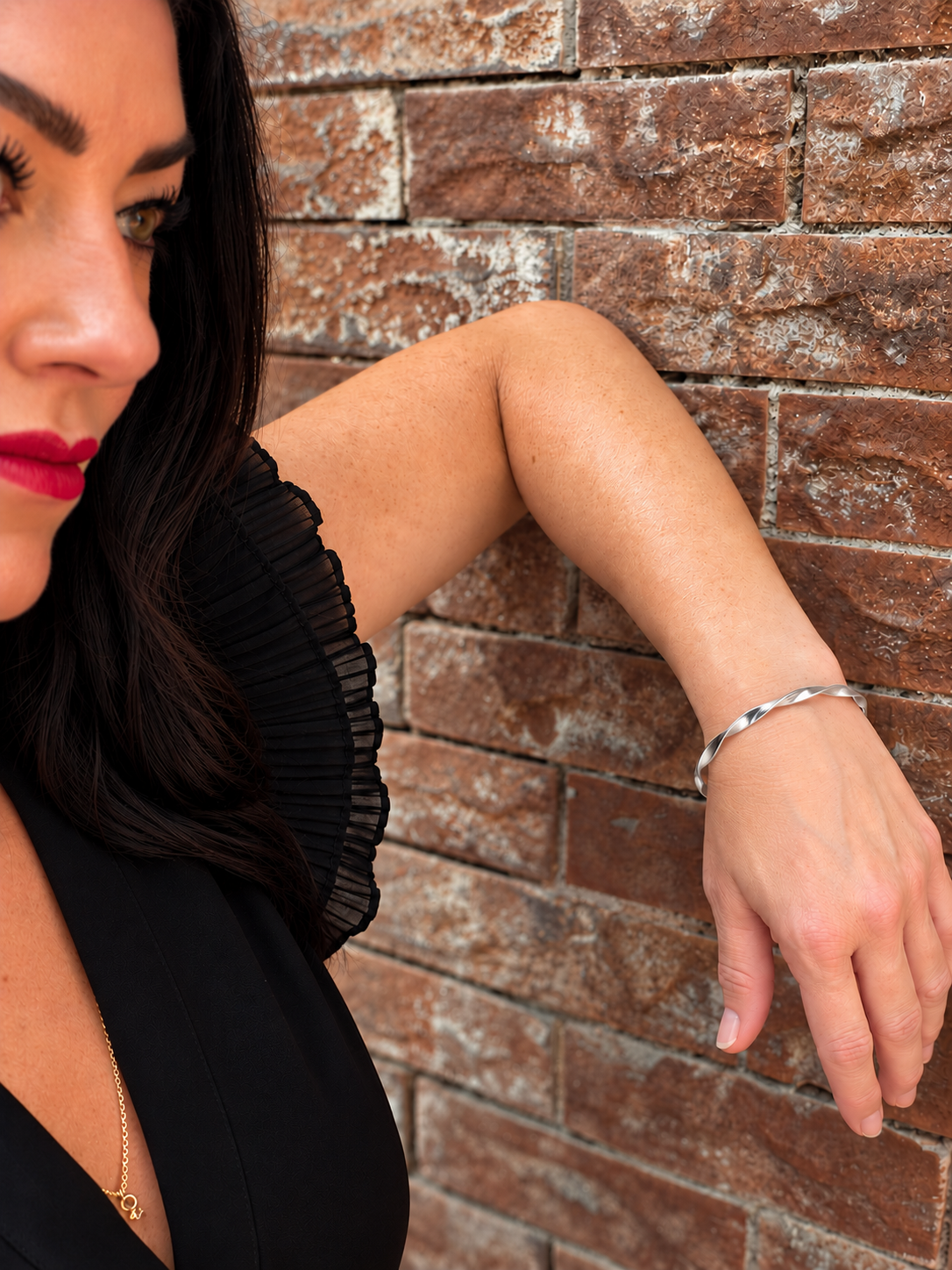 A woman wearing a polished silver bangle bracelet, leaning against a rustic brown brick wall.