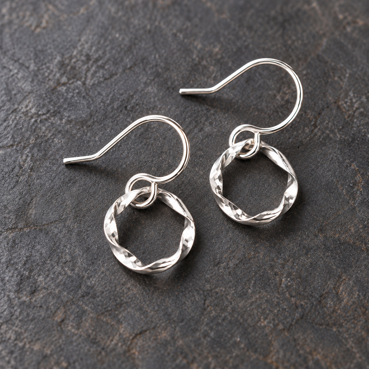 A pair of polished argentium silver twisted hoop earrings with fishhook backs, displayed on a dark textured stone surface.