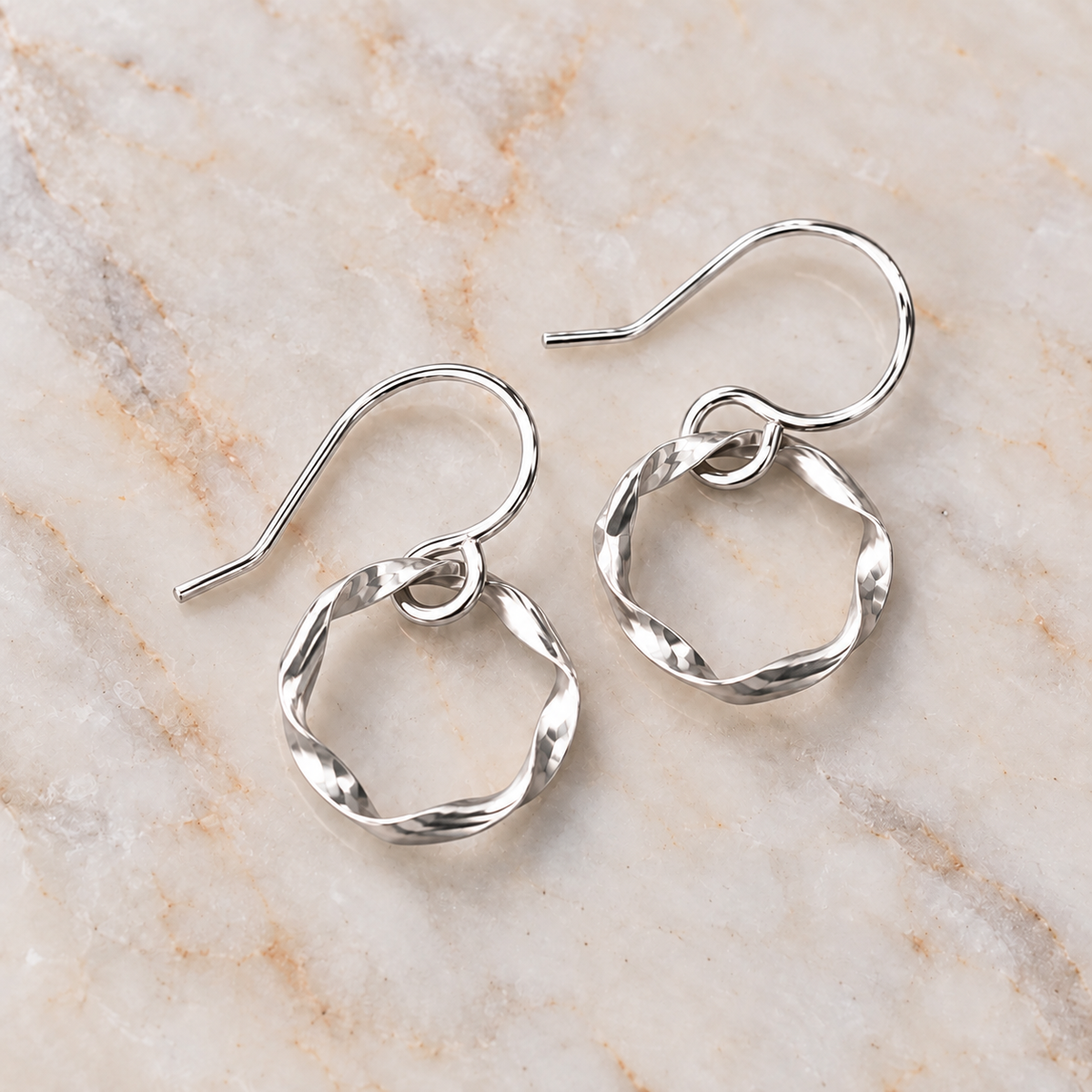A pair of silver dangle earrings with twisted open circular hoops and fishhook wires, displayed on a light beige marble surface.