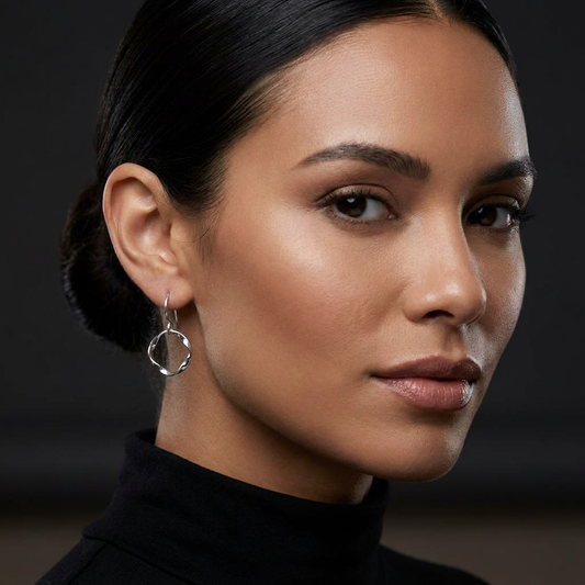 A close-up portrait of a woman with dark hair pulled back, wearing a single silver hoop earring with a small dangling circular accent. She is wearing Lepa Jewelry's Silver Strata Day Hoops in the polished option