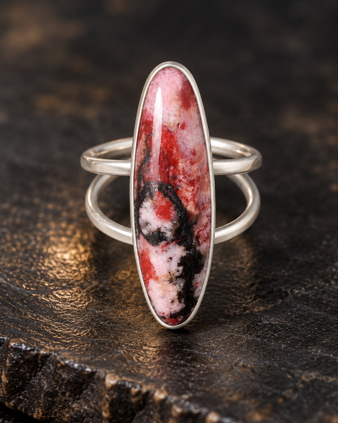 Front view of The Sola Ring by Lepa Jewelry, highlighting its slender rhodonite stone with dramatic rose-red and black markings against a dark textured background.