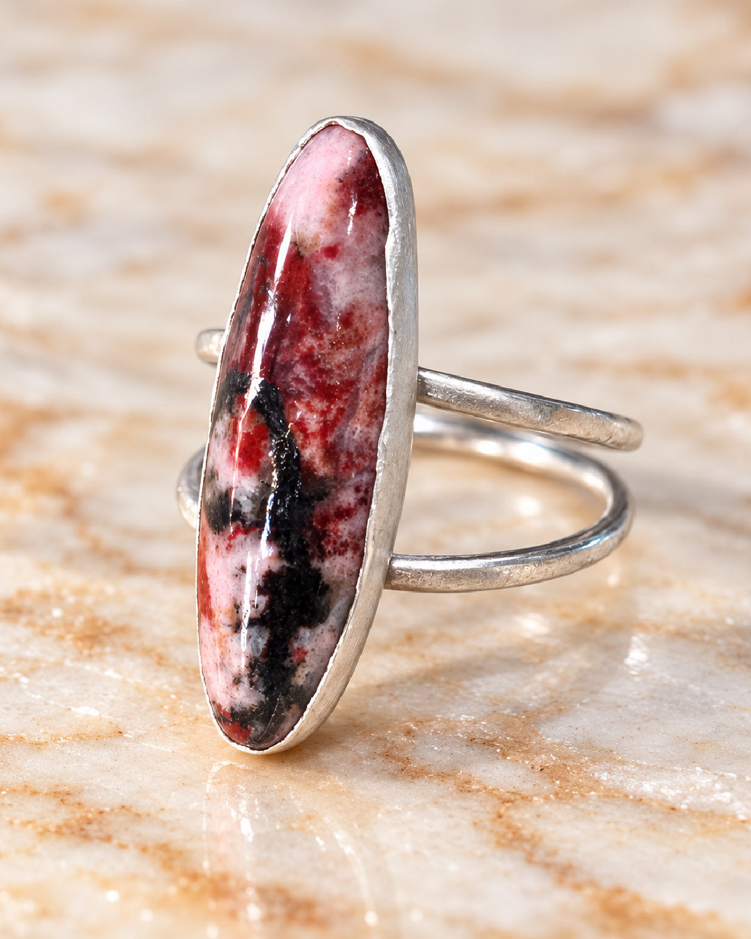 Three-quarter view of The Sola Ring showing the elongated rhodonite stone, fine silver bezel, and sculptural Argentium silver split-shank band on ivory marble.