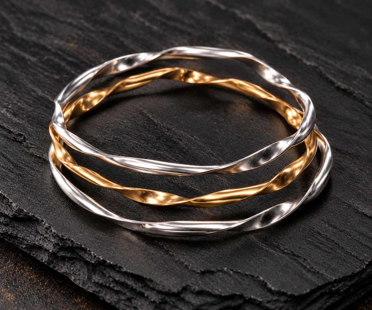 Three twisted metal bangles in Argentium silver and gold-filled metals, arranged in a layered circular stack on a dark textured stone surface.